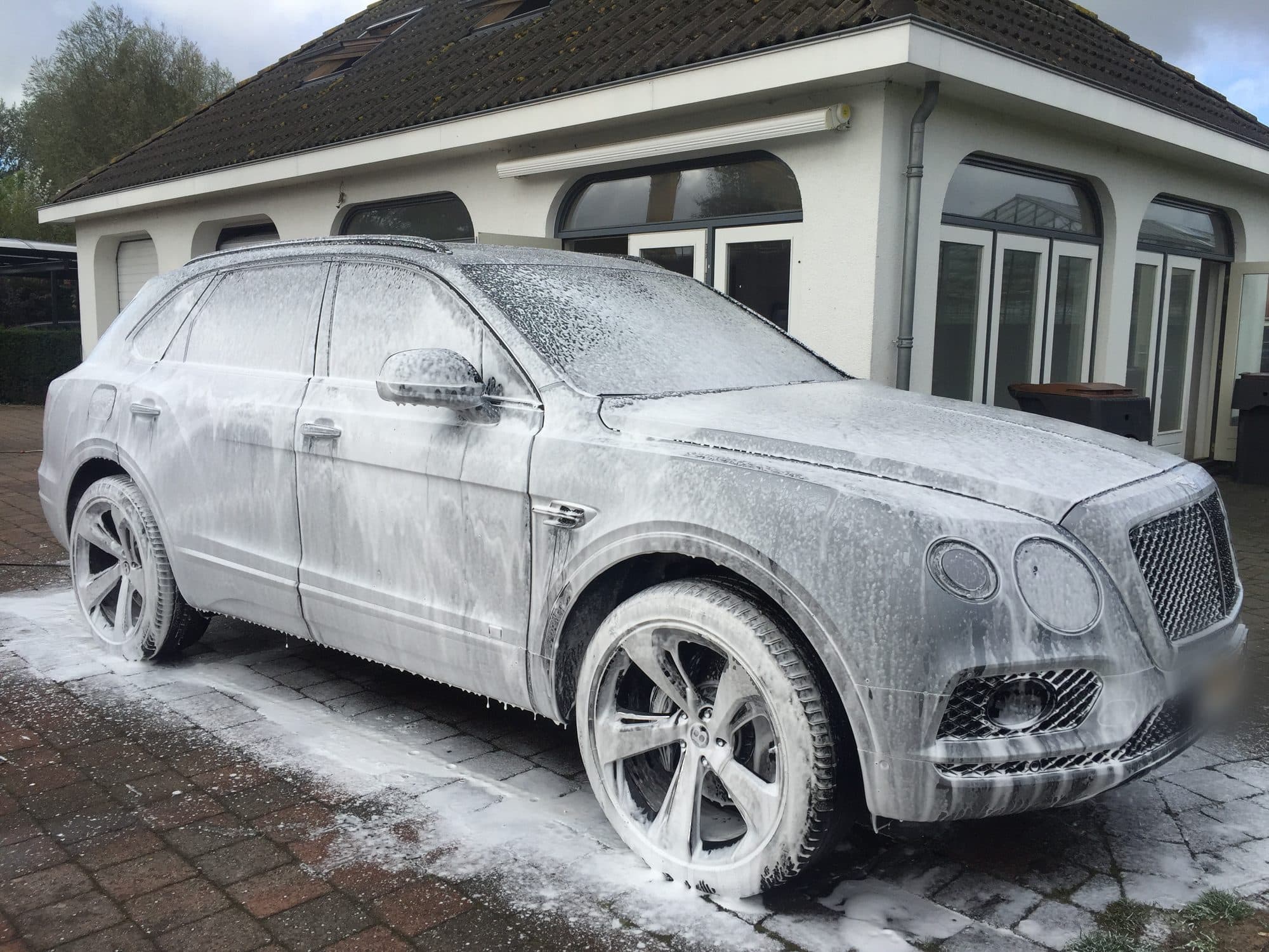 Een donkere luxeauto onder een laag wasschuim op een oprit, klaar om afgespoeld te worden.