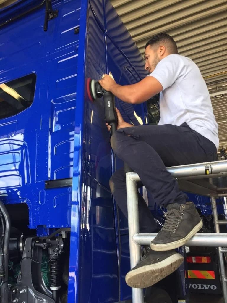 Een monteur zit op een rolsteiger en polijst met een machine de flank van een blauwe Volvo-truckcabine, binnen in een hal met een geribd dak.