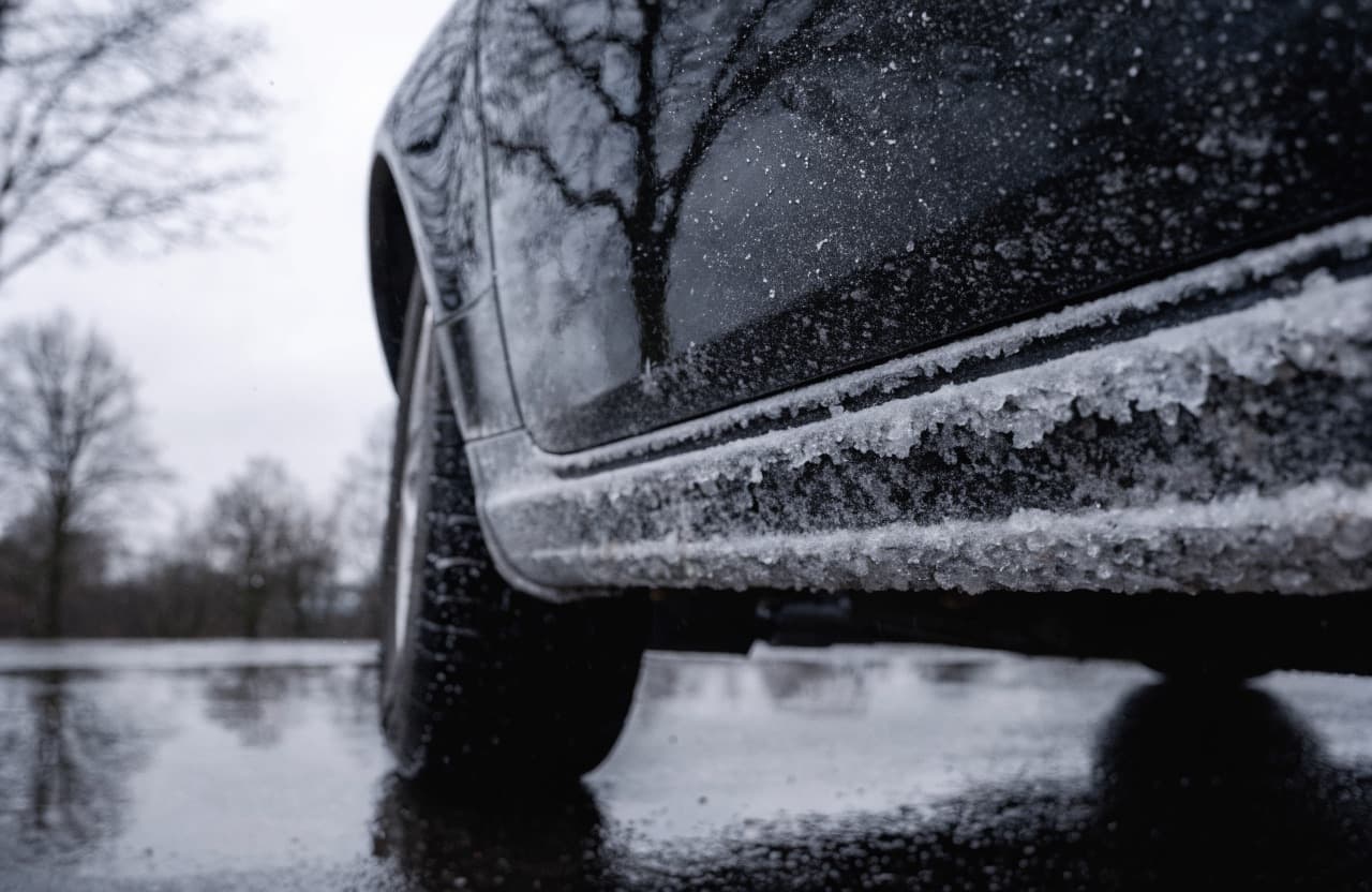 De onderkant en dorpel van een donkere auto zitten dik onder de opgedroogde witte zoutaanslag na een winterrit, met nat wegdek eronder.