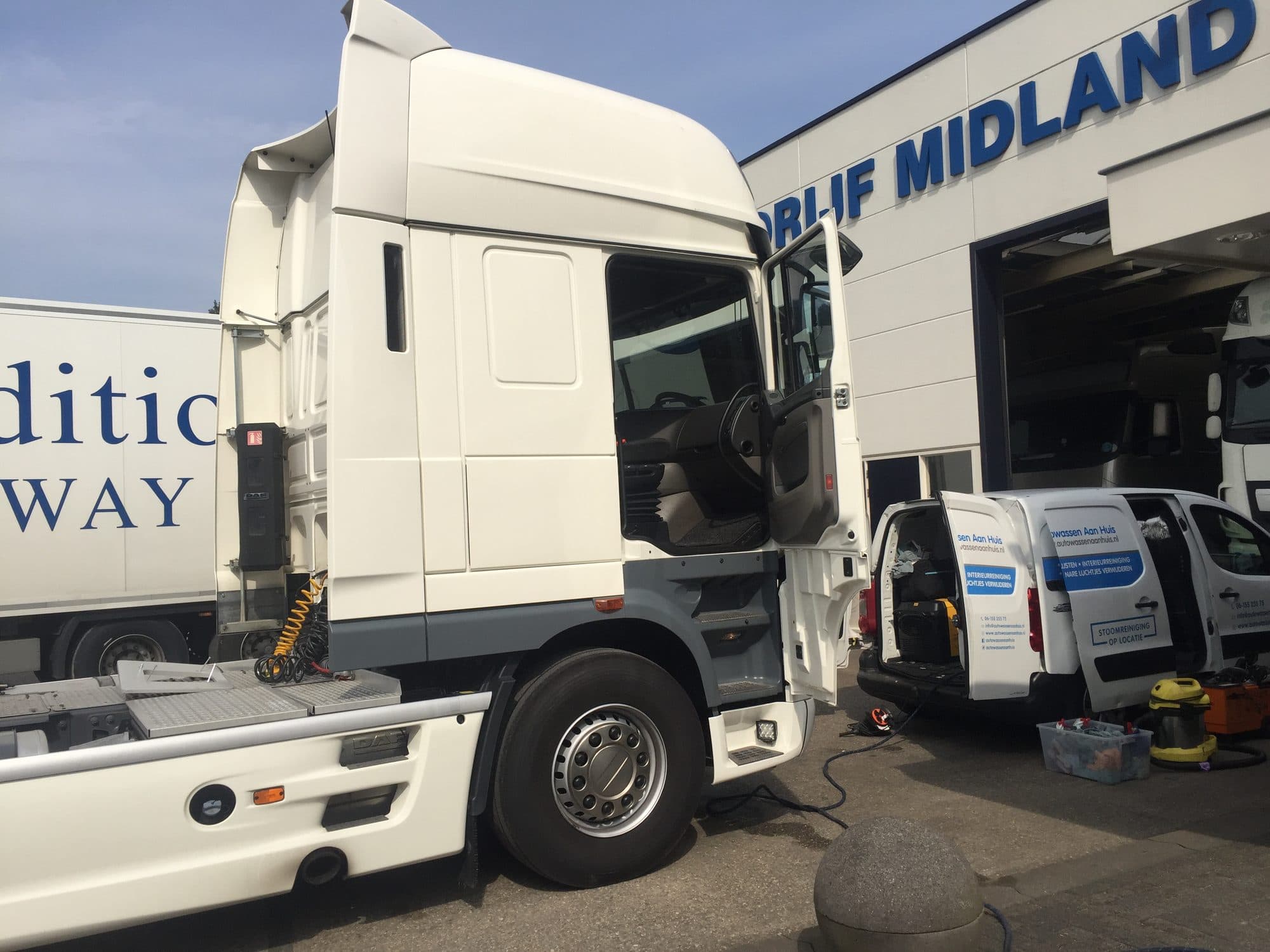 Witte trekker met open cabinedeur op een bedrijfsterrein, met daarnaast een bestelbus met reinigingsapparatuur en slangen die uitstaan.