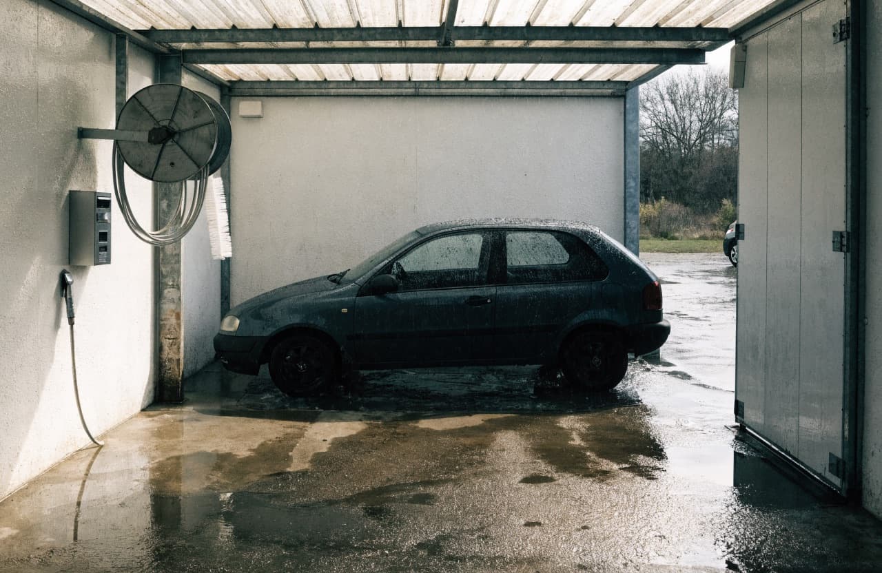 Een overdekte wasbox met muntautomaat aan de wand, een hogedruklans op een slanghaspel en een schuimborstel in de houder, met een natte auto in de box.