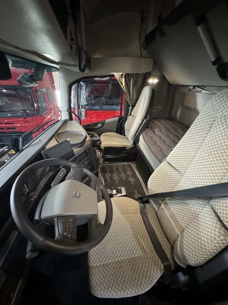 Het interieur van een Volvo-truckcabine na reiniging: lichte stoelen en slaapbank zonder vlekken, een schoongemaakt dashboard en een stofvrije vloermat, met door de voorruit een rode vrachtwagen zichtbaar.