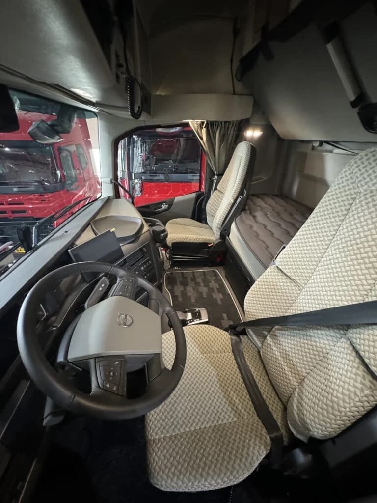 Het interieur van een Volvo-truckcabine na reiniging: lichte stoelen en slaapbank zonder vlekken, een schoongemaakt dashboard en een stofvrije vloermat, met door de voorruit een rode vrachtwagen zichtbaar.