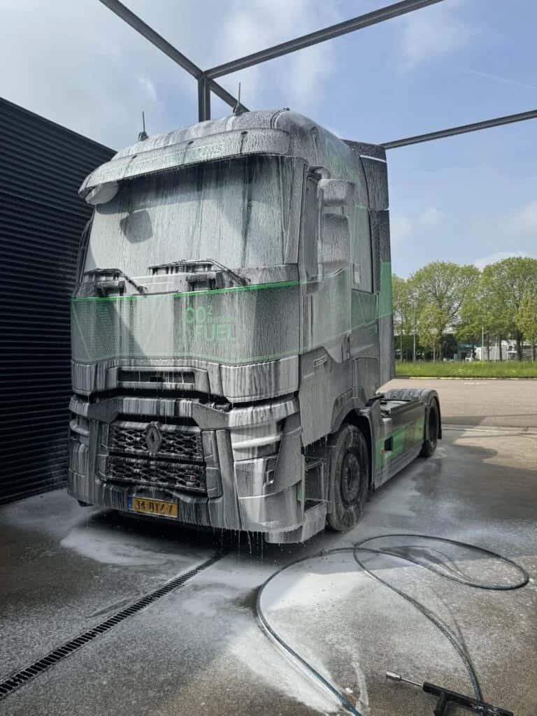 Een Renault-trekker met groene belettering staat volledig onder het schuim op een verharde vloer; in de voorgrond loopt een afvoergoot waar het waswater in wegloopt, met de hogedrukslang ernaast.