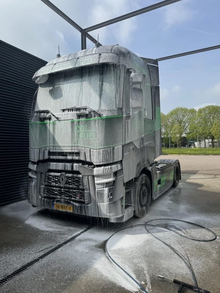 Een Renault-trekker met groene belettering staat volledig onder het schuim op een verharde vloer; in de voorgrond loopt een afvoergoot waar het waswater in wegloopt, met de hogedrukslang ernaast.